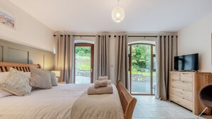 Een slaapkamer met een groot bed, een televisie op een houten dressoir en uitzicht op groen door de deur.