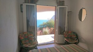 A room with a bed, two chairs, a sliding glass door leading to a balcony with a view of the sea and vegetation. - Villa Tarco, sleeping facilities, directly on the beach, 300 m from the beach (Conca)