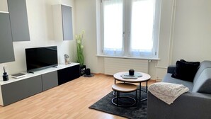 Un salon moderne avec un canapé gris, un téléviseur posé sur un meuble noir, une petite table ronde avec une tasse et une plante en pot.