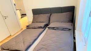 Un lit avec une tête de lit sombre et une literie grise, dans une pièce dotée d’un plancher en bois et d’une porte blanche.