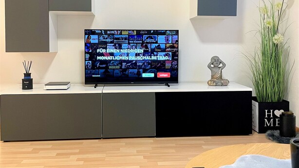 Un salon moderne avec un téléviseur à écran plat fixé au mur, une table basse en bois, une télécommande et une plante décorative.