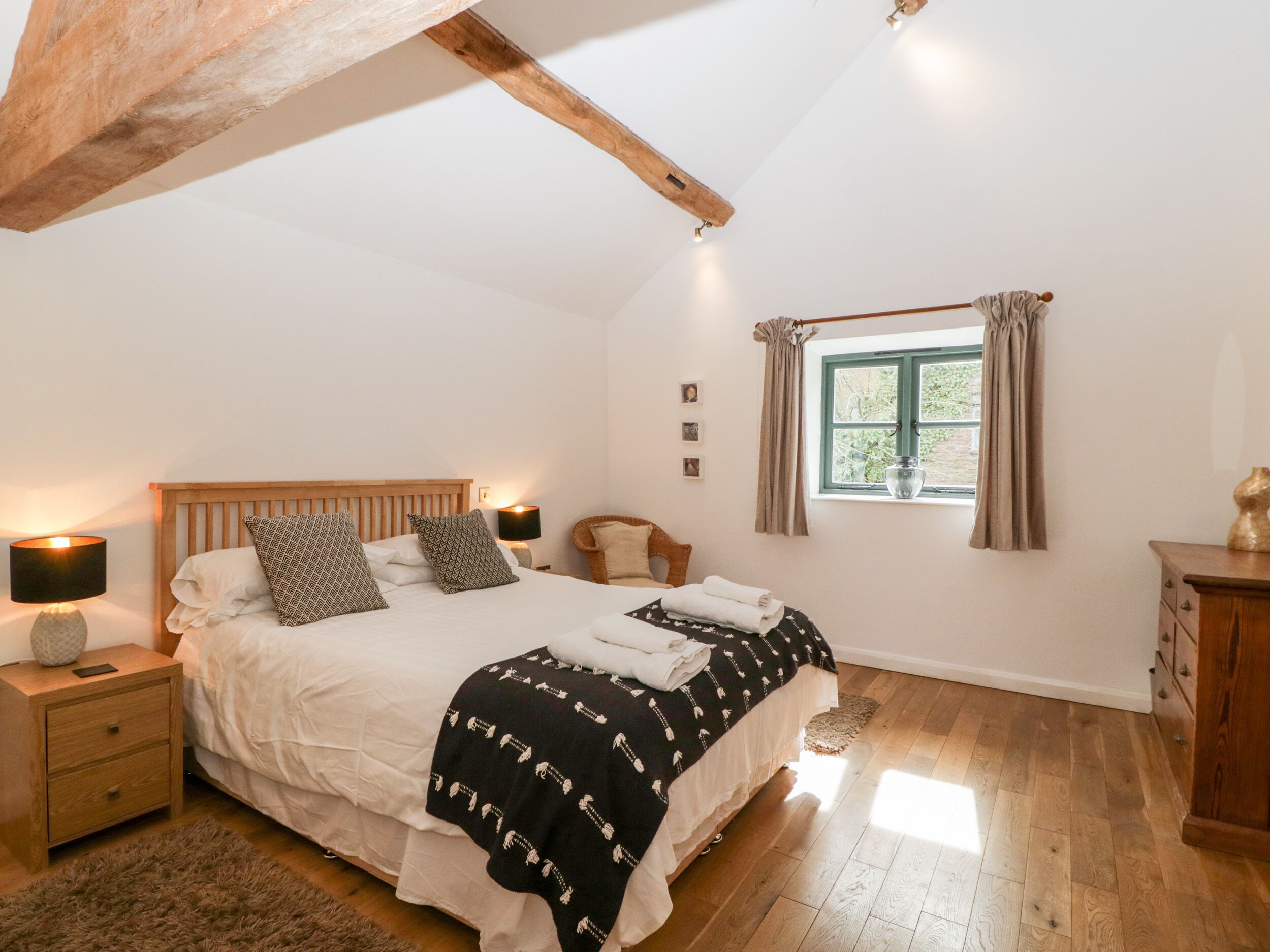 A bedroom with a wooden bed, a nightstand, a lamp, a window with curtains, and a wooden dresser.