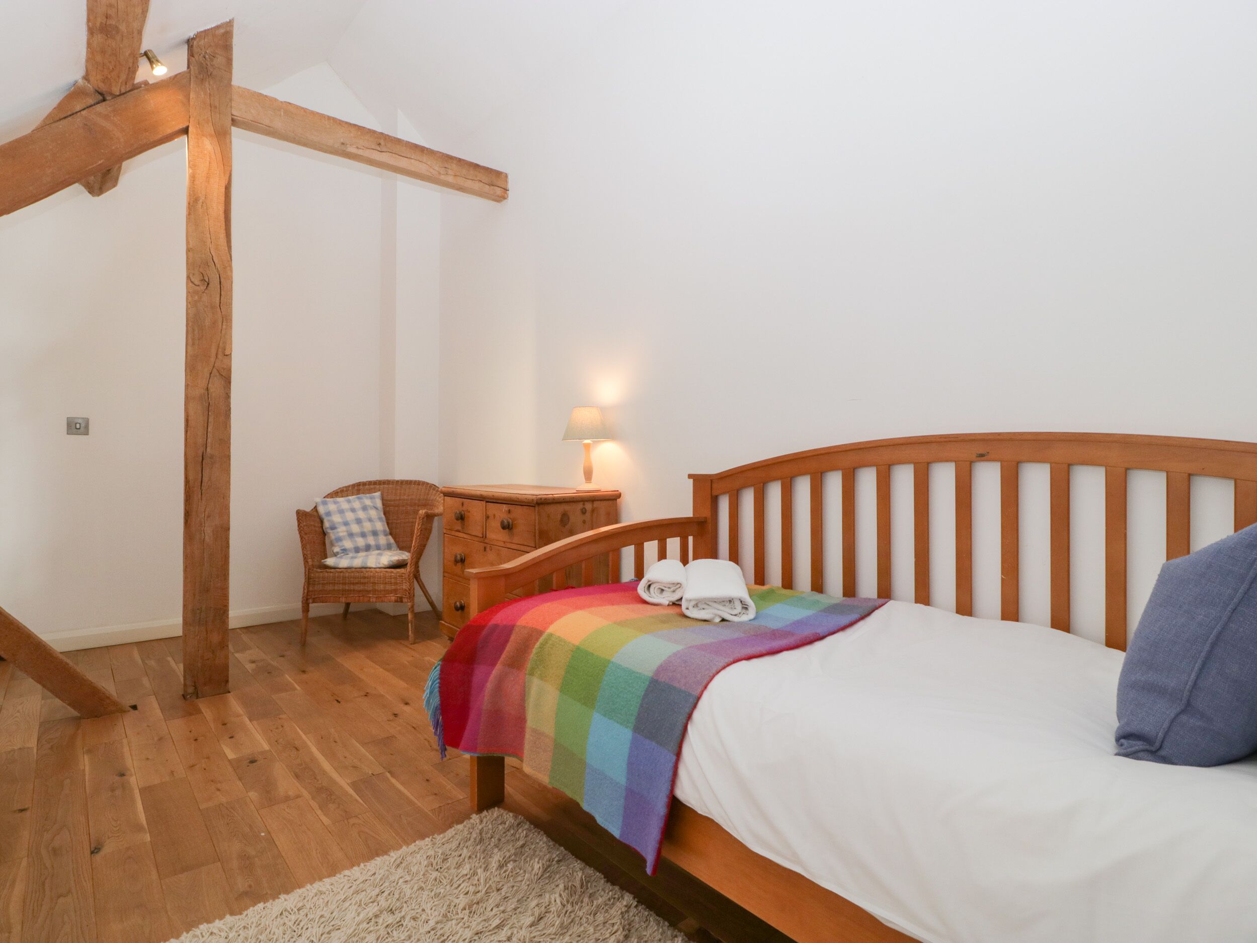 A bedroom with a wooden bed, a nightstand with a lamp, a wicker chair, and a wooden ceiling beam.