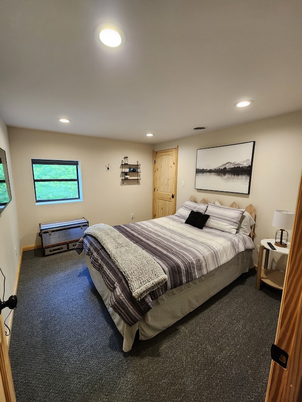 A bedroom with a large bed, a window, a wooden door, a shelf with items, and a painting on the wall. - Busse's Roost Cozy Mountain Home
(Cosby)