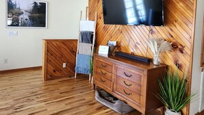 Ein geräumiger Raum mit Holzbalkendecke, einem an der Wand montierten Flachbildfernseher, einem Holzkasten mit Schubladen und einem großen Fenster, das für natürliches Licht sorgt.