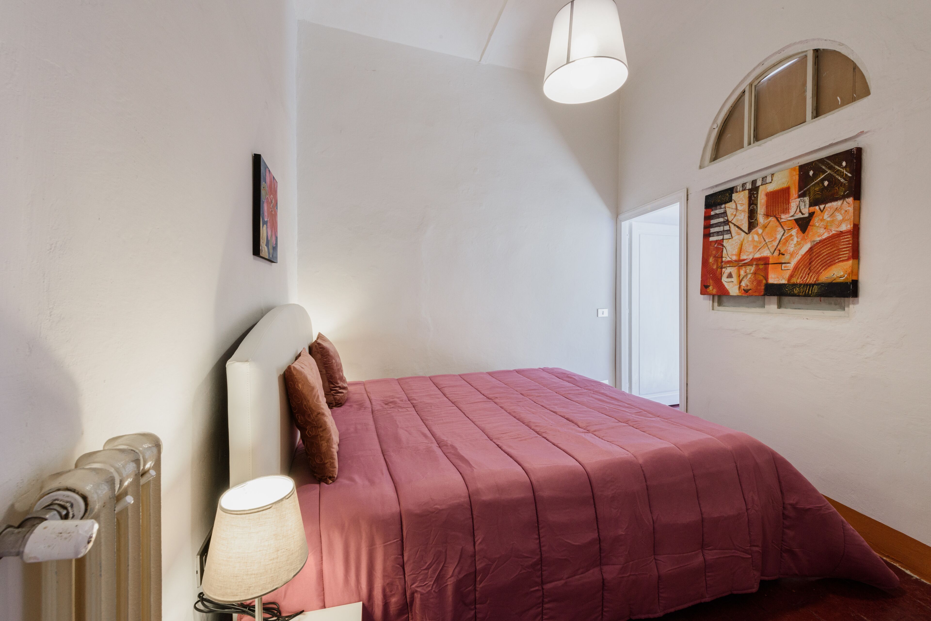 A bedroom with a large bed, a radiator, a lamp, and two paintings on the walls.