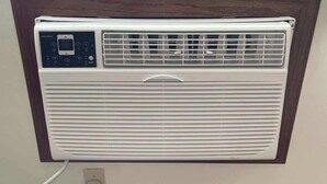 Aire acondicionado fijado a la pared, con panel de control, cable de alimentación y una ventana encima.