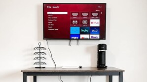 Moderni huone, jossa on seinälle asennettu televisio, jossa näkyy Roku-käyttöliittymä, kahvinkeitin pöydällä ja kaksi jakkaraa.
