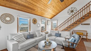 A spacious living room with a wooden ceiling, a large window, a staircase, and a cozy seating area. - Torchsong: Oceanfront, Direct Beach Access, Dogs OK (Nags Head)
