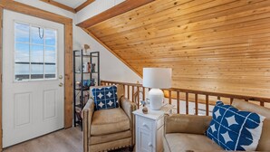 A cozy room with a wooden ceiling, a white door, a bookshelf, a chair, a sofa, and a nightstand with a lamp. - Torchsong: Oceanfront, Direct Beach Access, Dogs OK (Nags Head)