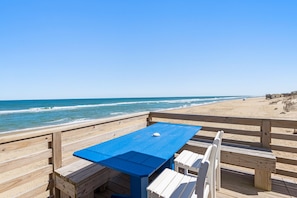 Une terrasse en bois avec une table bleue et des bancs blancs, donnant sur une plage avec l’océan en arrière-plan.
