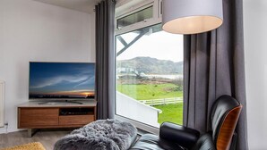 Ein modernes Wohnzimmer mit großem Fenster, einem Flachbildfernseher, einem Ledersofa und einem flauschigen Hocker.