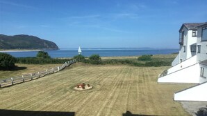 Ein grünes Rasenstück mit einem kleinen, runden Blumenbeet, einem weißen Zaun und Blick auf das Meer und die Hügel im Hintergrund.