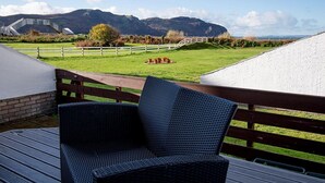 Eine Terrasse mit Korbstühlen, einem Holzzaun und Blick auf ein Wiesfeld mit einem weißen Gebäude und Bergen im Hintergrund.
