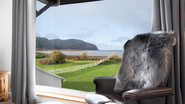 Ein Zimmer mit Blick auf eine Wiese und ein Gewässer, umzäumt von einem weißen Zaun, mit einem Sessel und einem Fellüberwurf.