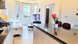 Une cuisine moderne équipée d’une cuisinière intégrée, un coin repas avec une table et des chaises blanches, et un téléviseur fixé au mur.