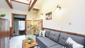 A cozy living room with a grey sofa, wooden beams, stone wall, and a small table with a plant.