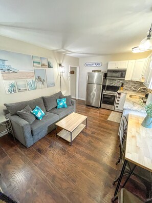 Un espacio habitable compacto con un sofá gris, una mesa de centro de madera y una cocina equipada con nevera, microondas y fogones.