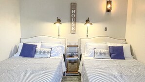 Ein Hotelzimmer mit zwei Betten, jeweils mit gestreifter Bettwäsche und blauen Kissen. An der Wand hängt ein Schild mit der Aufschrift 'HOUSE' und zwei Wandlampen.