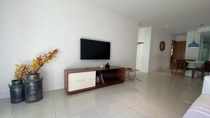 Een moderne woonkamer met een flatscreen televisie, een houten kast en een eethoek met een glazen tafel en stoelen.