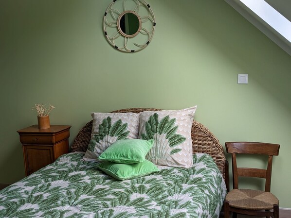 A bedroom with a bed, a green wall, a mirror, a wooden nightstand, a wicker chair, and a small table with a vase.