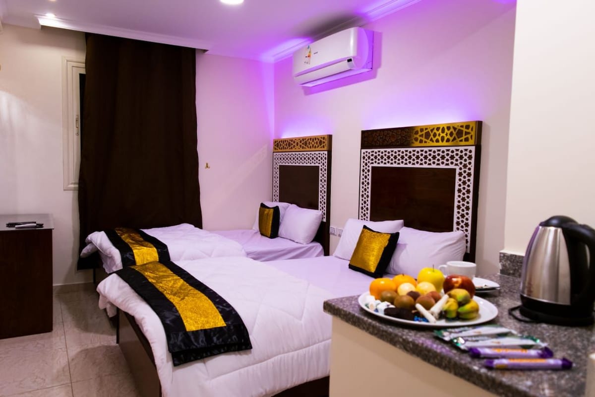 A hotel room with two beds, a small desk, and a breakfast bar with fruits and a kettle.
