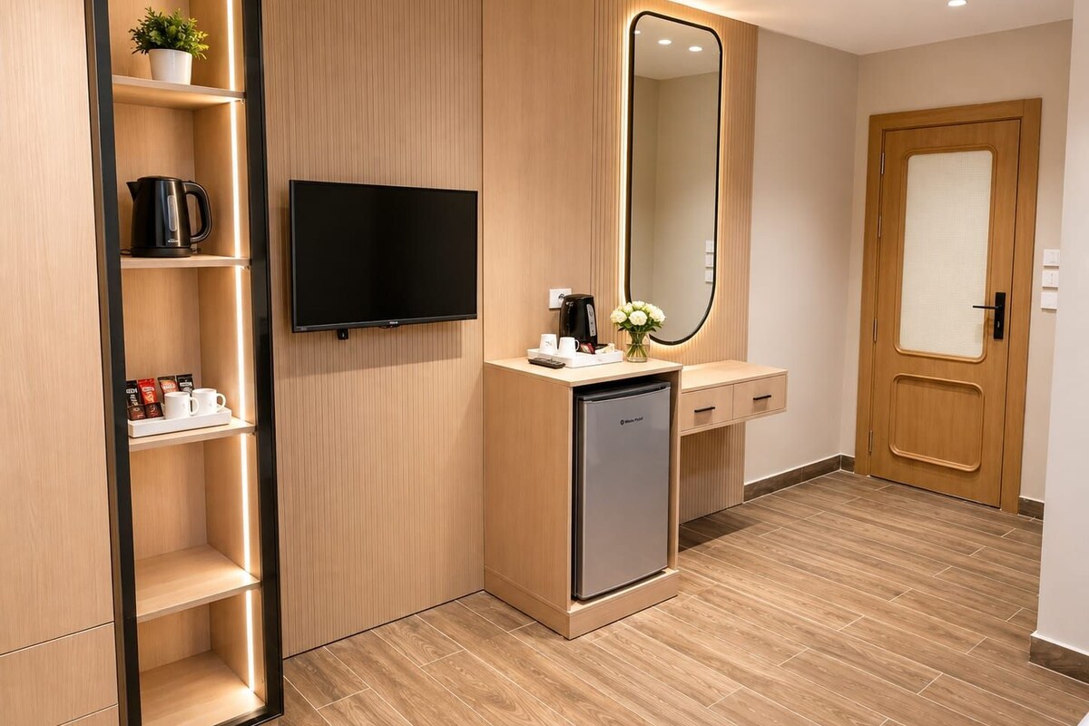 A modern hotel room with a flat-screen TV, a built-in coffee maker, a mini-fridge, a wooden door, and a large mirror.