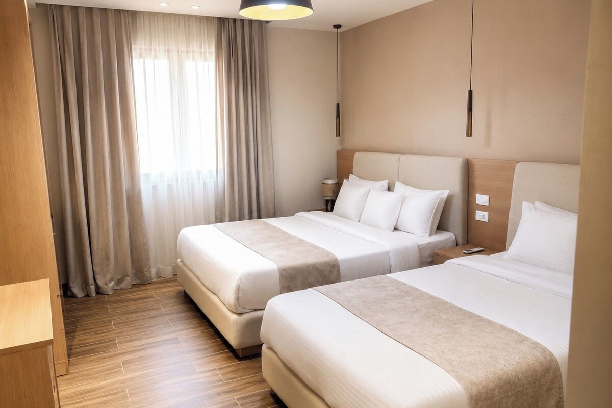 A hotel room with two beds, a wooden floor, a window with curtains, and a modern pendant light.