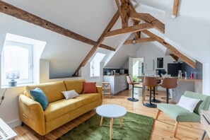 Un loft moderne avec des poutres en bois apparentes, un canapé jaune, un fauteuil vert et une cuisine à l’arrière-plan.