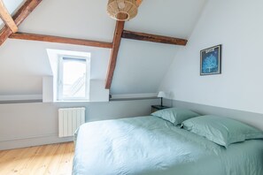 Une chambre avec un plafond en pente, une fenêtre, un lit avec une literie de couleur vert clair, une table de chevet avec une lampe et un tableau encadré au mur.