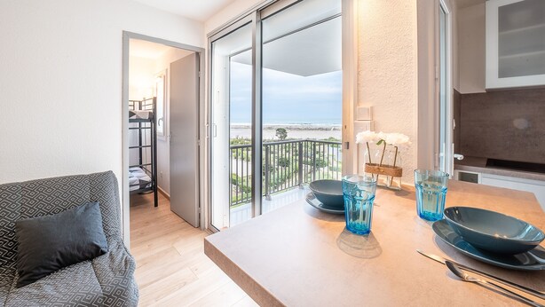 Un salon moderne avec un canapé, une table à manger agrémentée de verrerie bleue, et une vue sur la mer grâce à de grandes portes coulissantes.