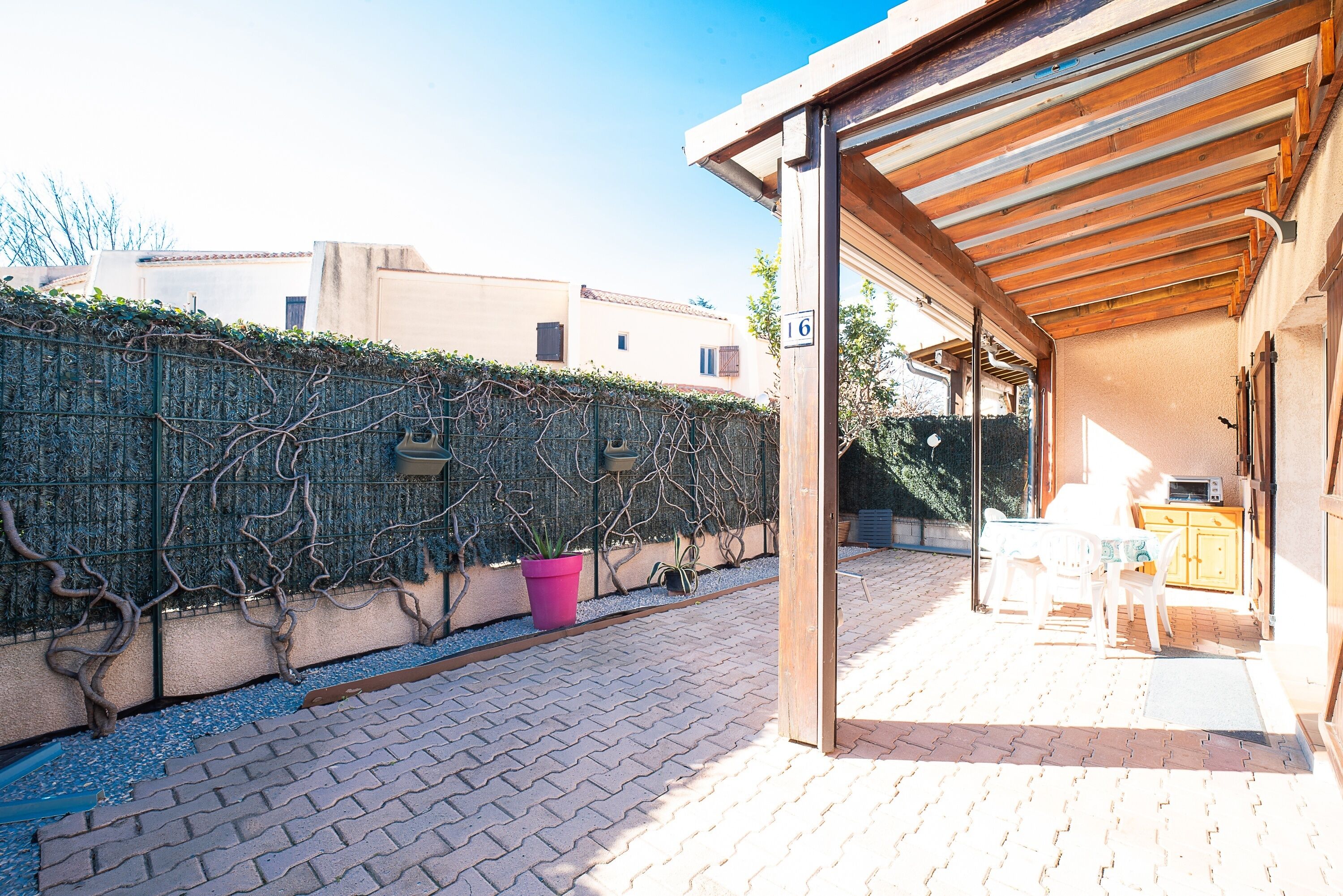 Un patio extérieur abrité, doté d’une pergola en bois, d’un chemin en briques et d’un petit espace détente avec une table et des chaises.