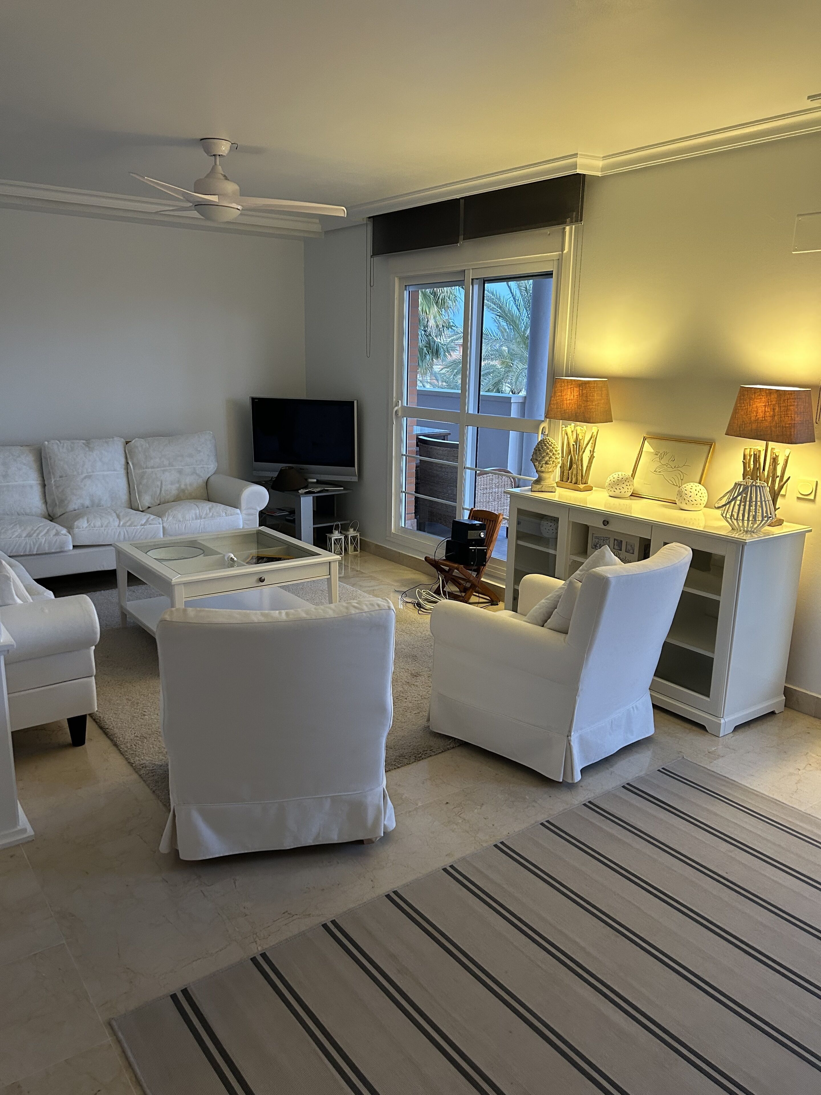 A modern living room with a white sofa, a coffee table, and a TV. There is a window with a view of palm trees outside.