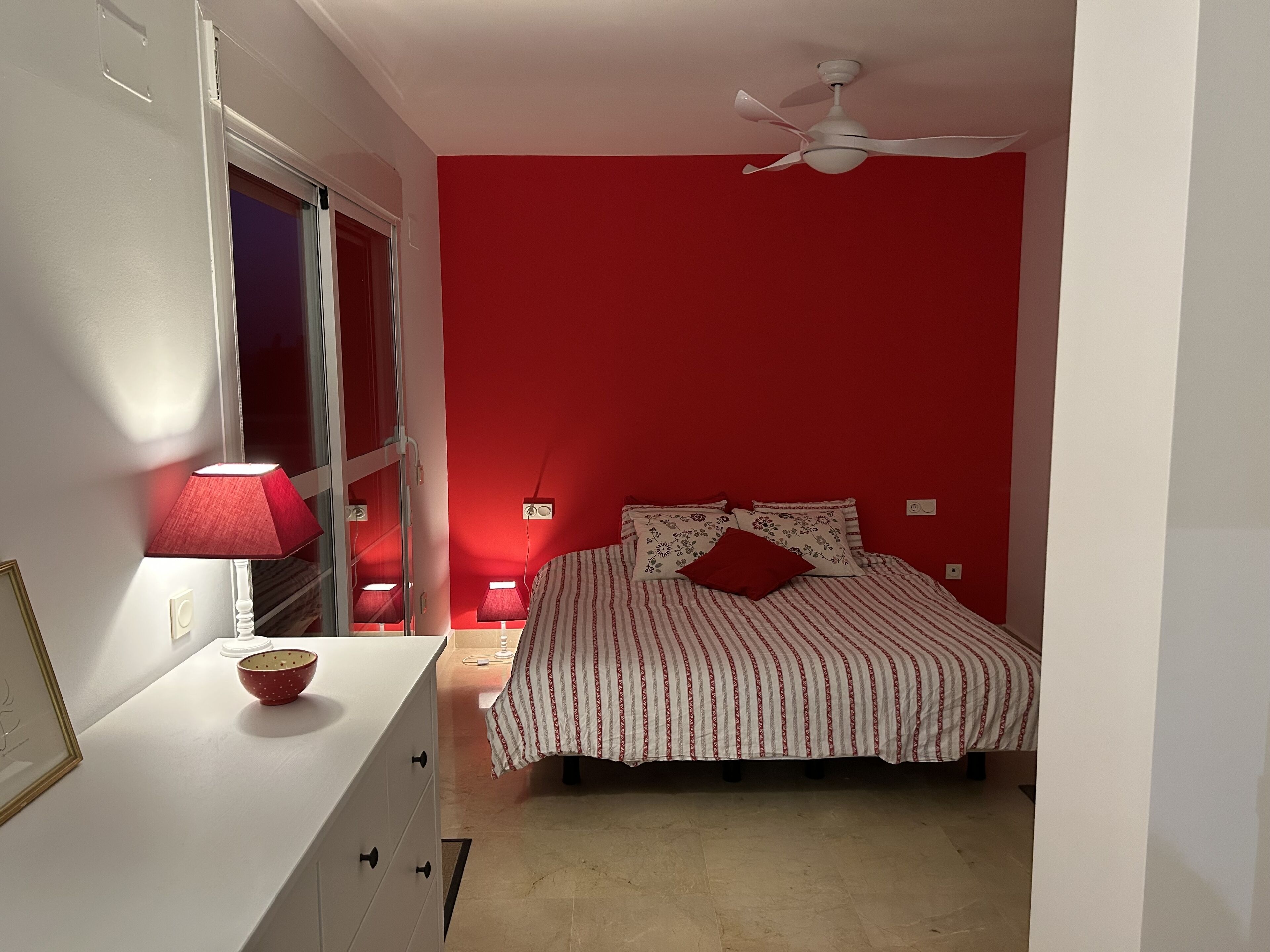 A bedroom with a bed, a ceiling fan, a red wall, and a window with a red lamp.