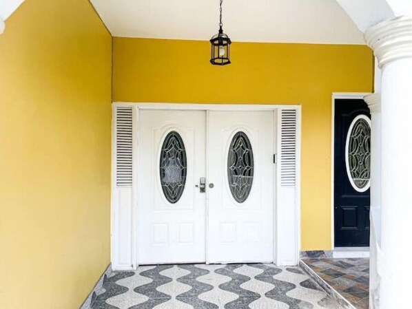 A yellow exterior wall with a white door featuring decorative glass panels and a black door with a similar design. - Exceptional 2Bdrms-Red Hassell Manor, Port Antonio (Port Antonio)