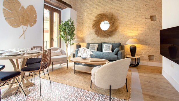 A cozy living room with a sofa, coffee table, and chairs, a dining table with plates and glasses, a large plant, and a round mirror on a brick wall. - Genteel Home Santas Patronas II (Seville)
