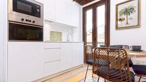 A compact kitchen with a built-in oven, microwave, and a small sink. There is a dining area with wicker chairs and a table, and a window with a view of buildings outside. - Genteel Home Santas Patronas III (Seville)