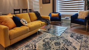 Un saló amb un sofà groc, una butaca blava, una taula de café de vidre i una catifes amb dibuixos.