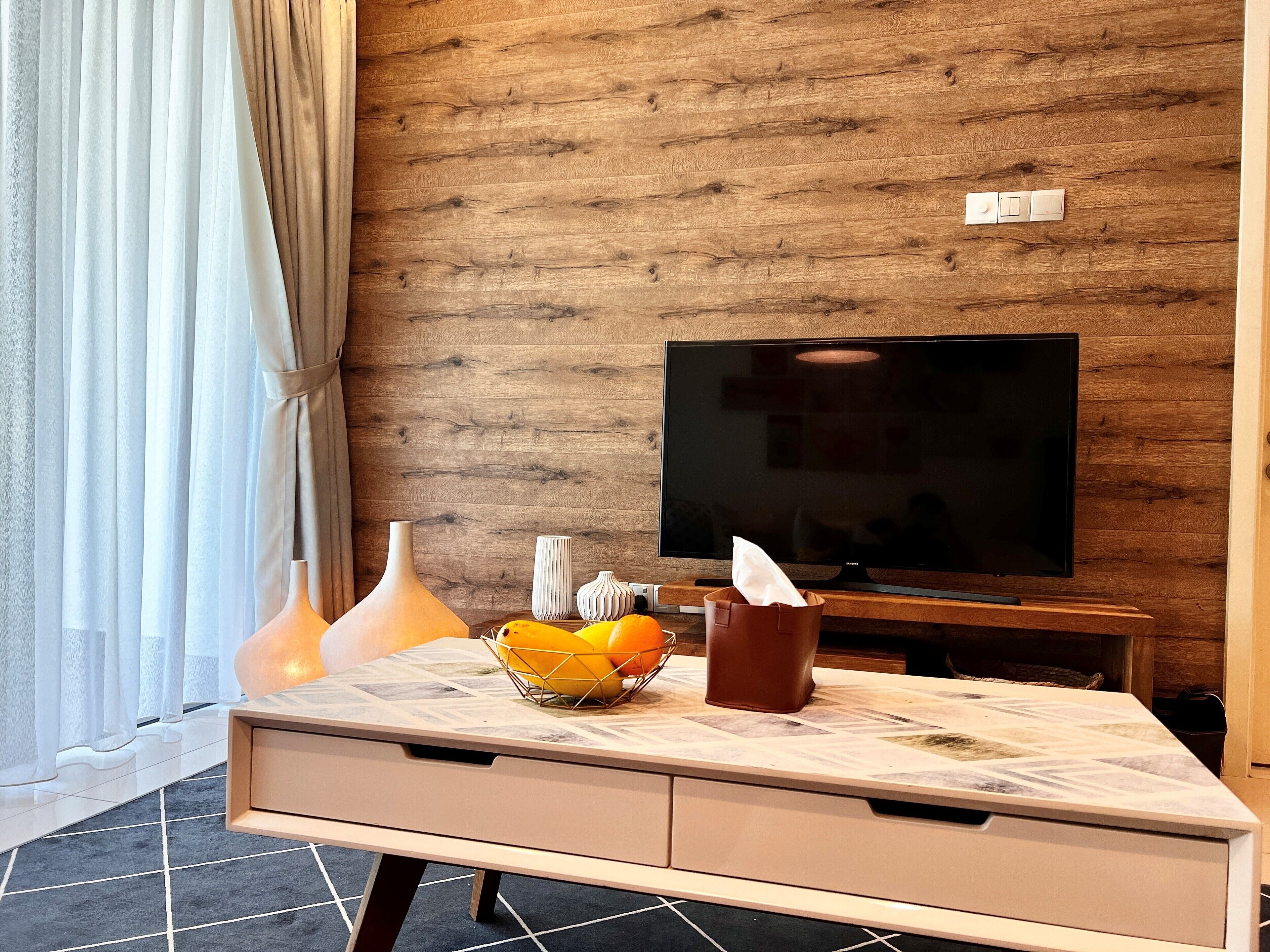 Een moderne woonkamer met een houten wand, een flatscreen televisie, een marmeren salontafel en een raam met dunne gordijnen.