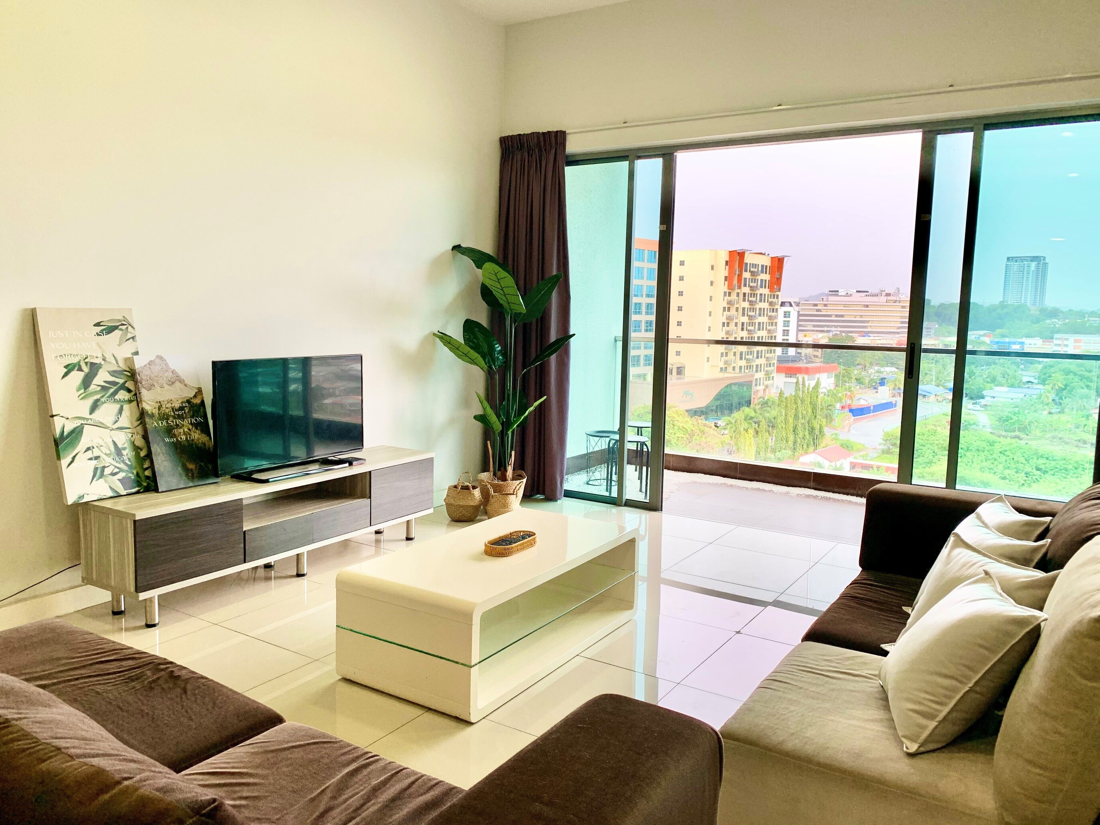 A modern living room with a flat-screen TV, a coffee table, and a sofa. There is a large window with a view of buildings and greenery outside.