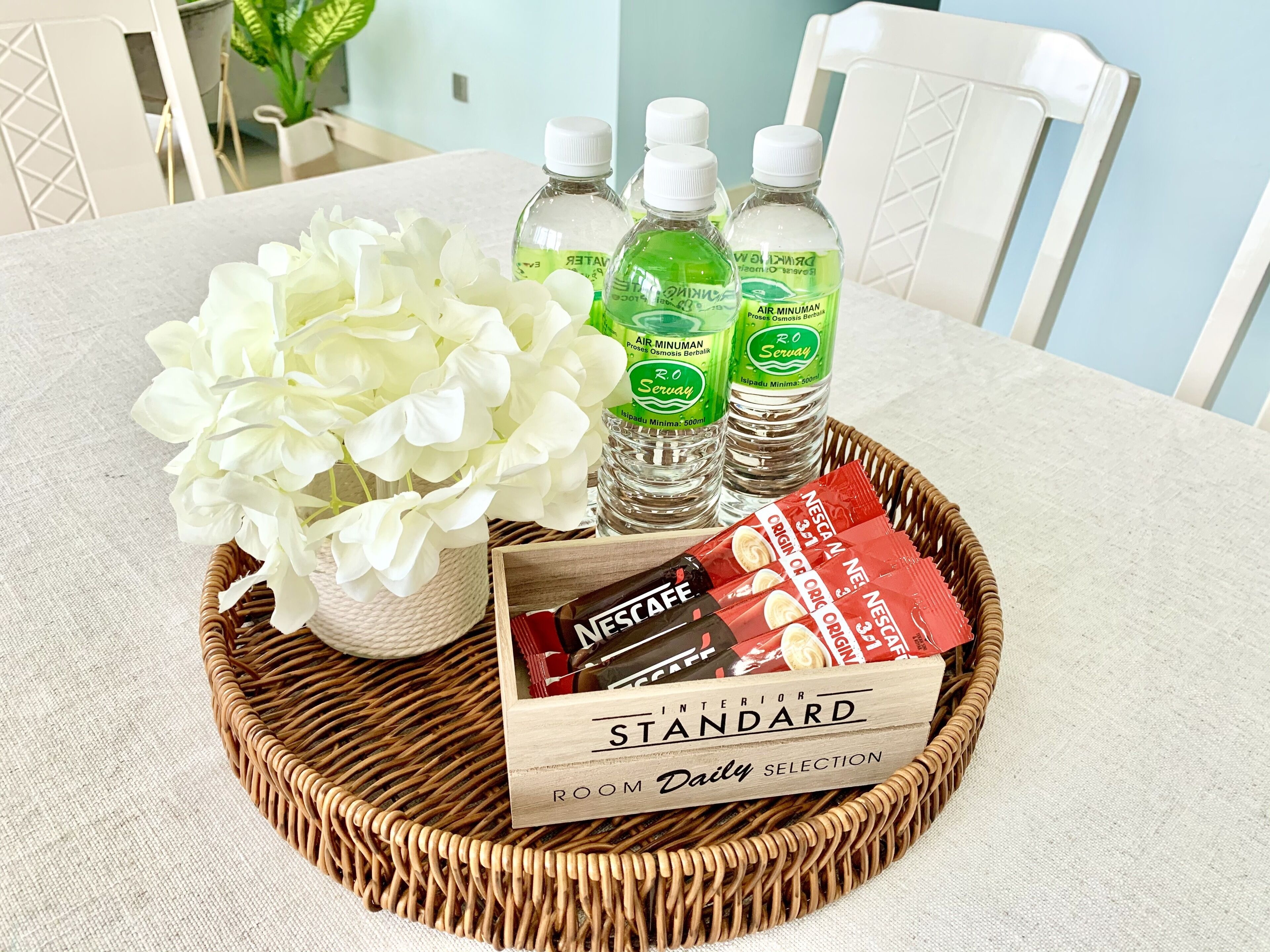 A wicker tray with a bouquet of white flowers, three bottles of water, and a box of Nescafé coffee on a table.