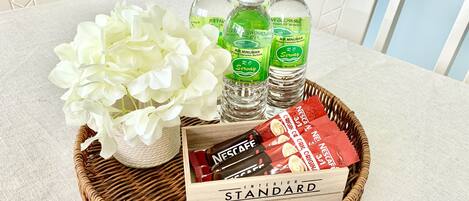 A wicker tray with a bouquet of white flowers, three bottles of water, and a box of Nescafé coffee on a table.