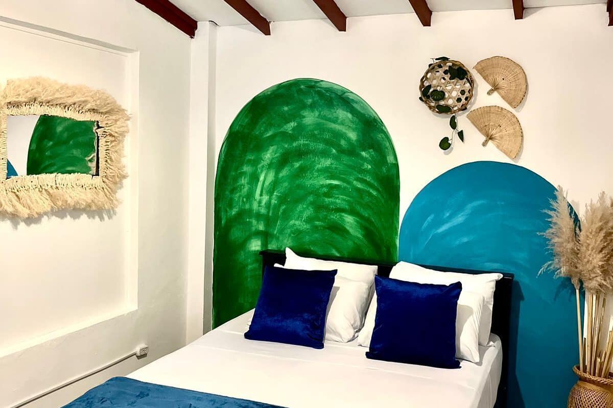 Quarto com cama, arco na parede verde e azul, luminária pendente e uma pequena planta.