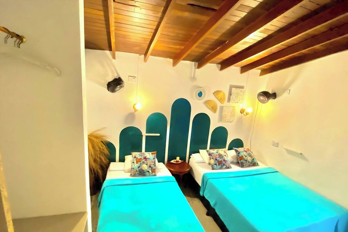 Quarto com duas camas, cada uma com colcha azul-turquesa e almofadas estampadas. O quarto possui vigas de madeira no teto e uma pequena mesa com abajur.