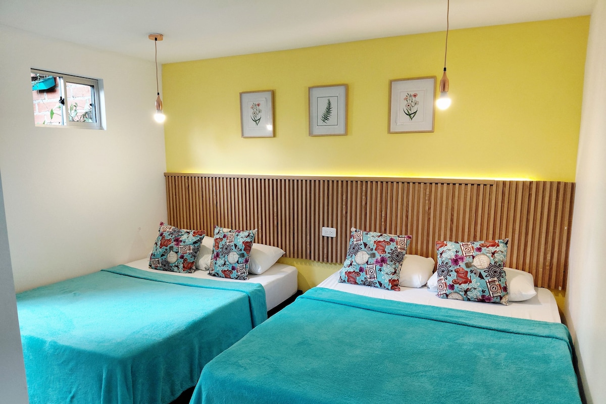 Habitación de hotel con dos camas, cada una con cubrecama color turquesa y almohadas estampadas con motivos florales, cabecero de madera con listones y tres láminas botánicas enmarcadas en la pared.