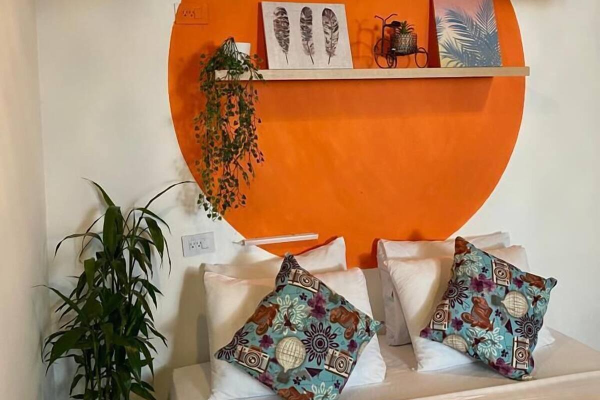 Quarto com parede laranja grande, cama com roupa de cama branca e almofadas estampadas, luminária pendente e prateleira com objetos decorativos.