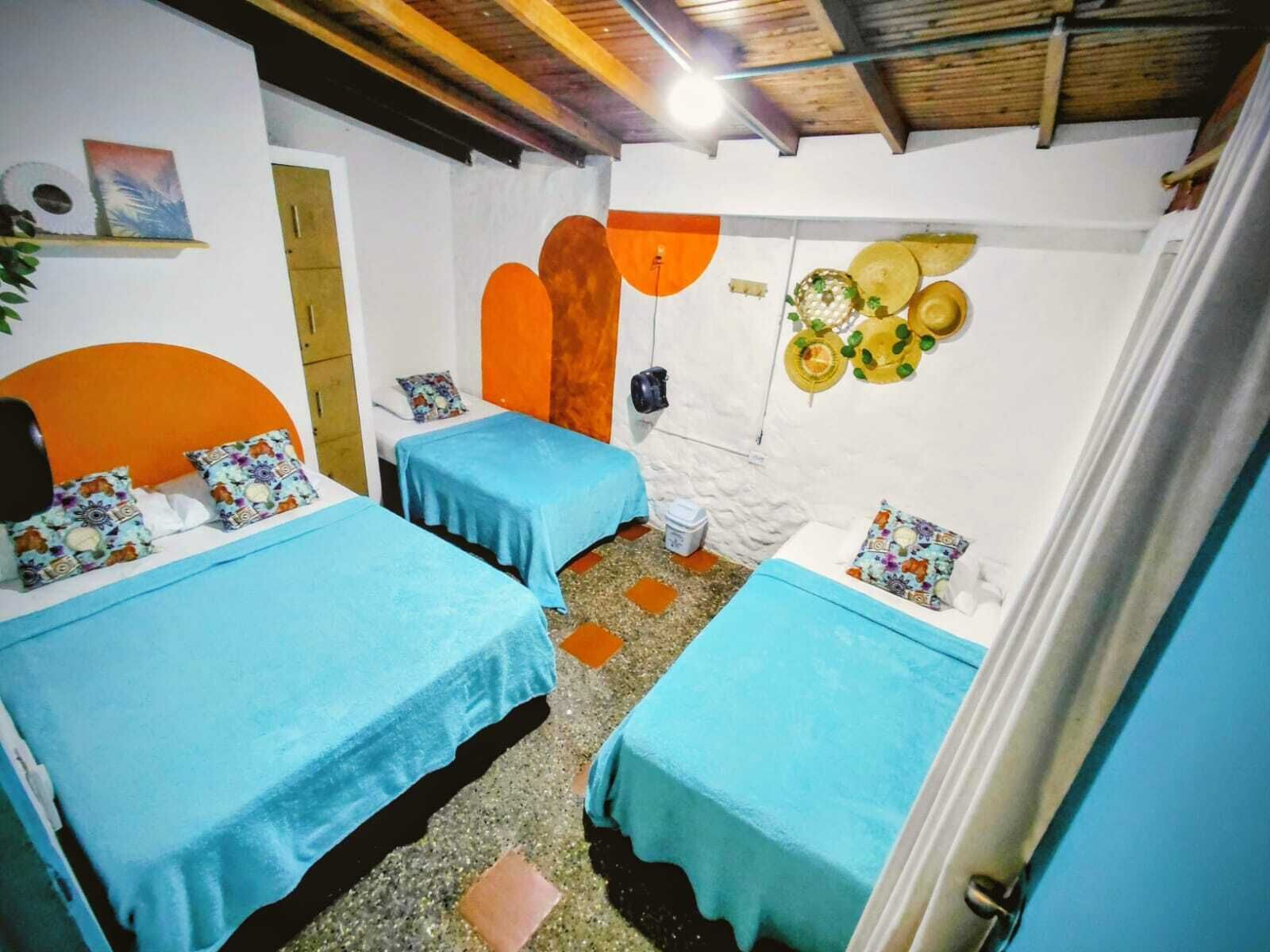 Habitación con dos camas, techo de madera, pared blanca decorada con adornos naranja y amarillo, y una pequeña mesa con un cubo.