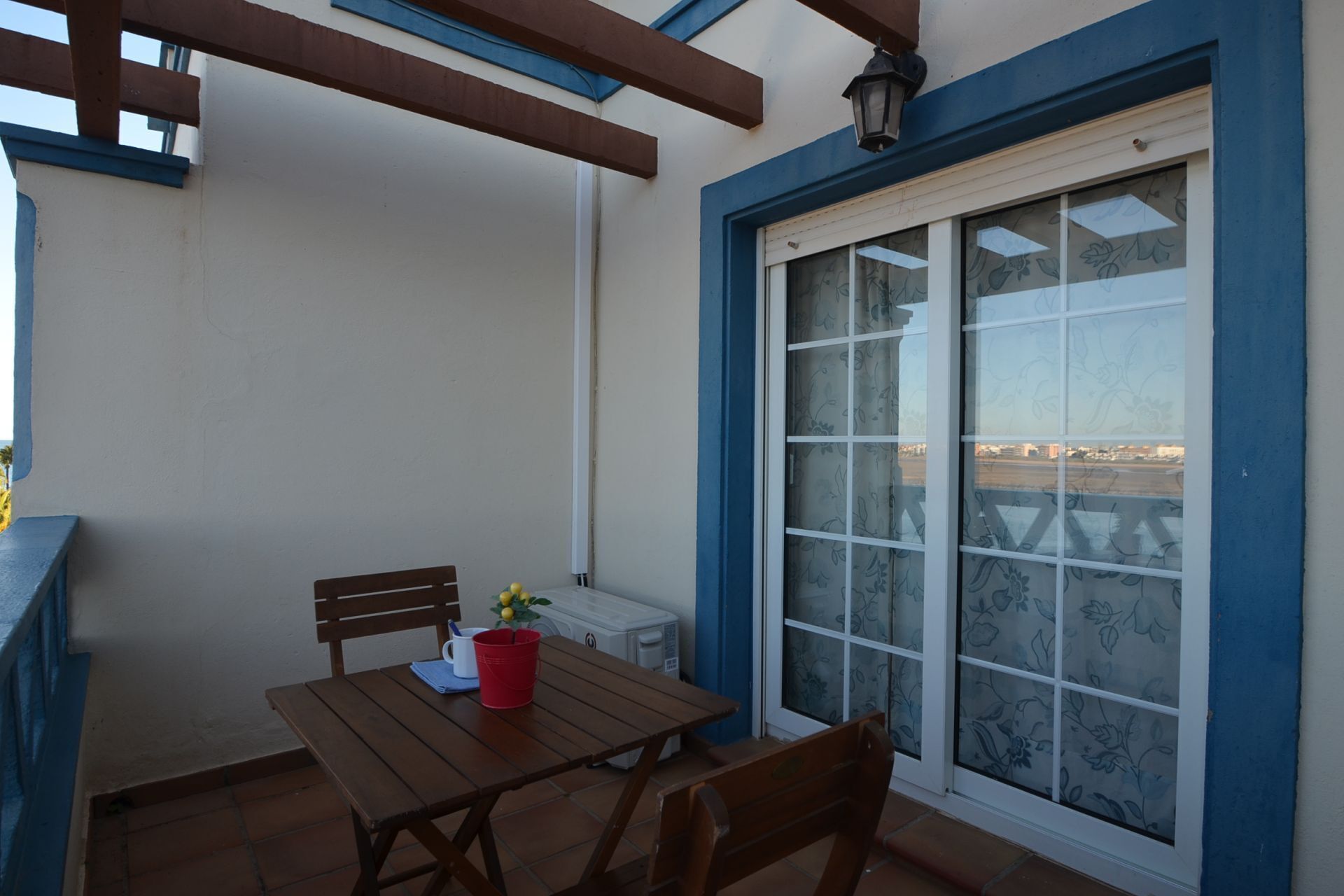 A balcony with a wooden table, chairs, a red bucket, and a small potted plant.