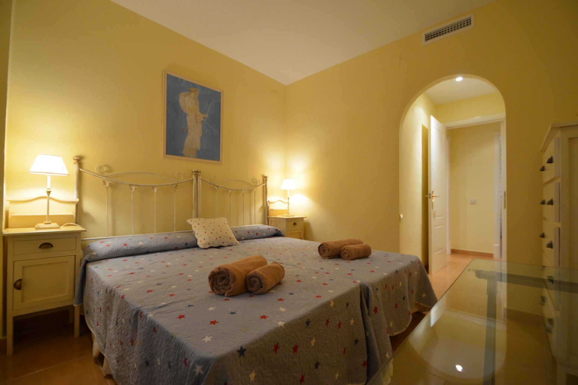A bedroom with a bed, two bedside tables, a nightstand, a painting on the wall, and a door leading to another room.