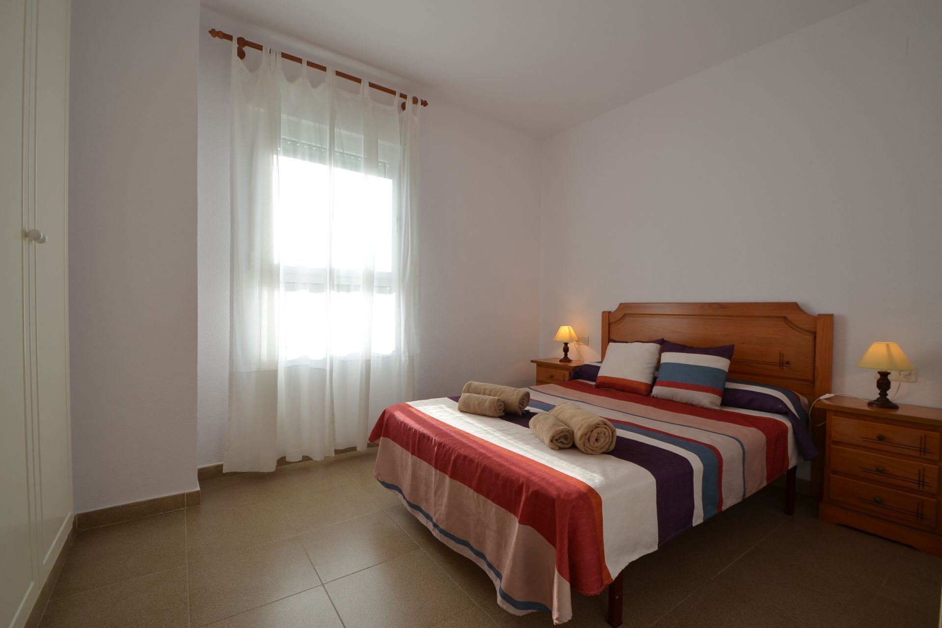 A bedroom with a bed, two bedside tables with lamps, a window with sheer curtains, and a door.